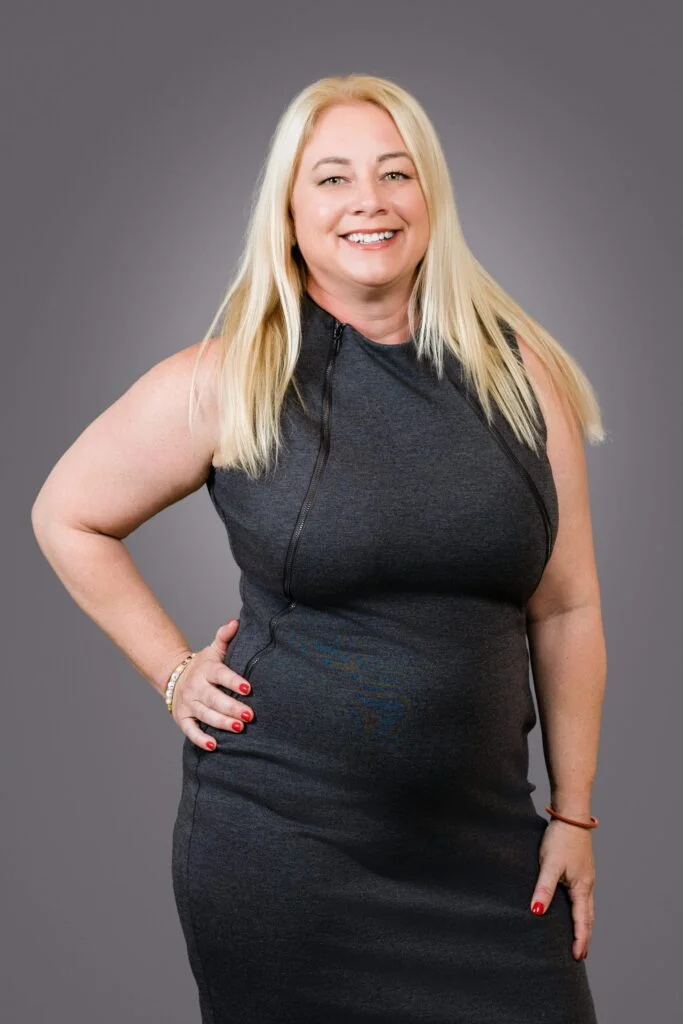 Portrait of Jenny Pelletier, Senior Executive Administrator at Hyde Park Capital, wearing a charcoal dress, smiling confidently in front of a gray studio backdrop.